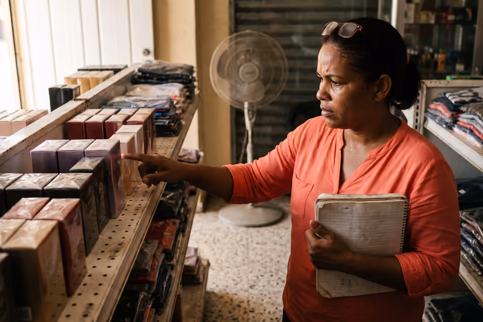 Dueña de una tienda de variedades contando productos en el estante con una libreta apoyada en el antebrazo.