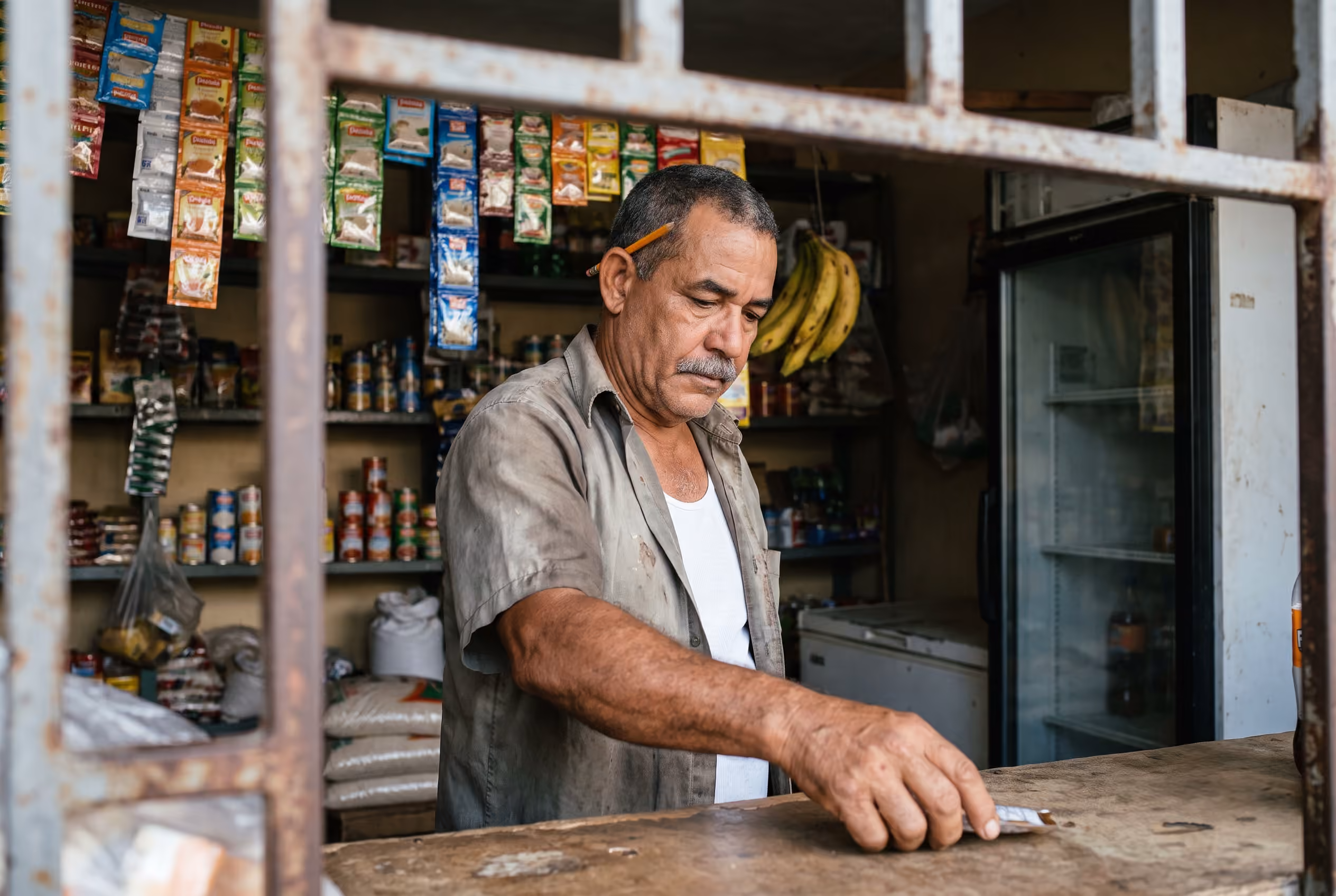 Dueño de un colmado dominicano contando billetes sobre el mostrador, con la nevera de refrescos al fondo.