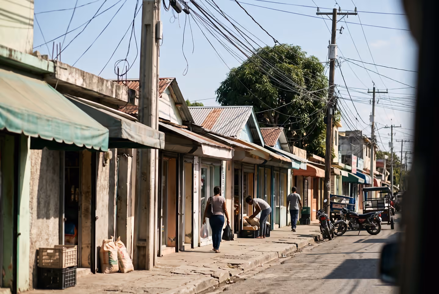 Una cuadra de comercios pequeños dominicanos vista a lo largo de la acera: toldos de colores distintos, cables aéreos cruzando la calle, motores parqueados y unas pocas personas de espaldas en una mañana de día laborable.